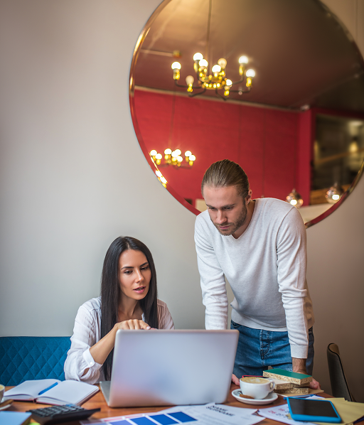 Man and woman collaborating and looking at a laptop on a table with papers, notebook, and coffee cups in a modern room with a round mirror reflecting chandeliers.